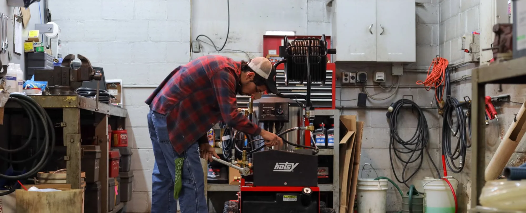 A service technician in a dealership shop working on a Hotsy hot-water pressure washer, tools and parts shelves around him.